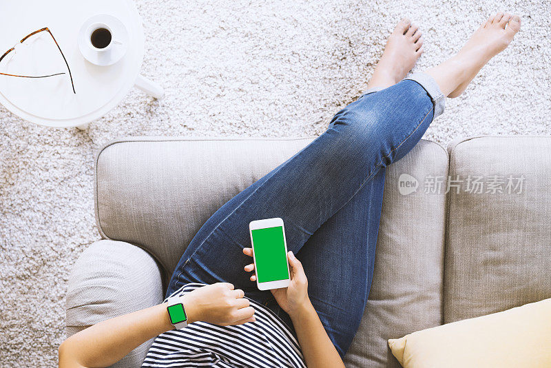 坐在沙发上用绿色屏幕智能手表和手机的女人。