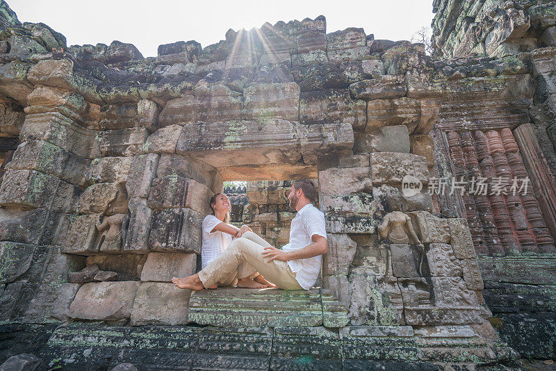 年轻夫妇凝视着古庙，坐在石墙的窗户上环顾四周，放松地花时间呼吸这一切，日落时分的阳光