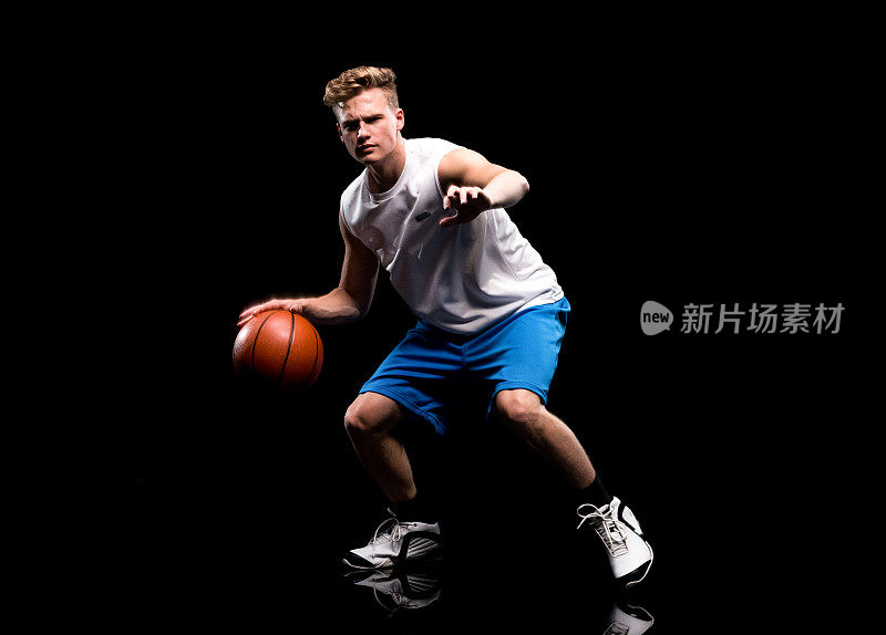 白人少年无袖蹲在黑色背景前，穿着运动鞋，拿着篮球和使用运动球