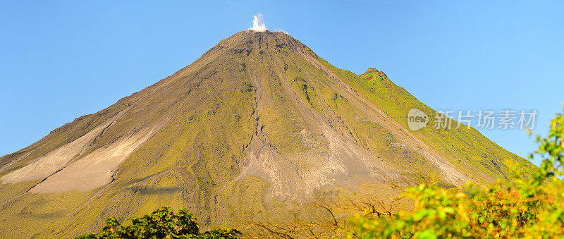 阿里纳斯火山在晴朗的天空下冒着蒸汽，两侧是郁郁葱葱的绿色植物，顶部蒸汽上升