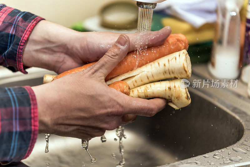 一个男人在水槽里彻底清洗胡萝卜和防风草的照片。