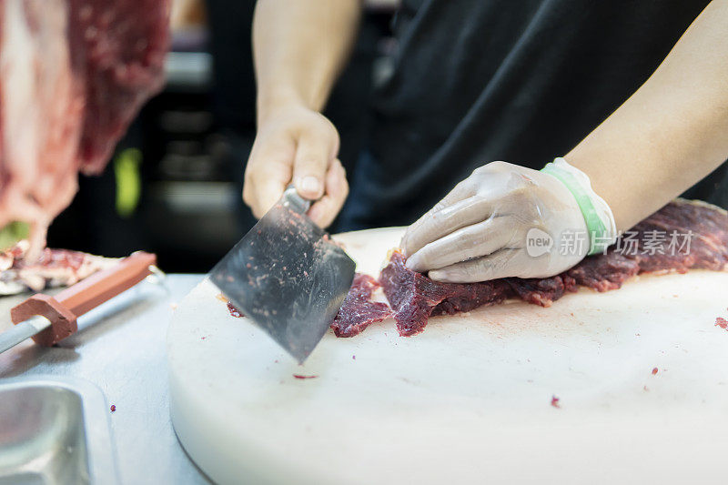 在一家牛肉火锅餐厅的开放式厨房里，厨师在挂着的切碎的生肉后面用刀切牛肉