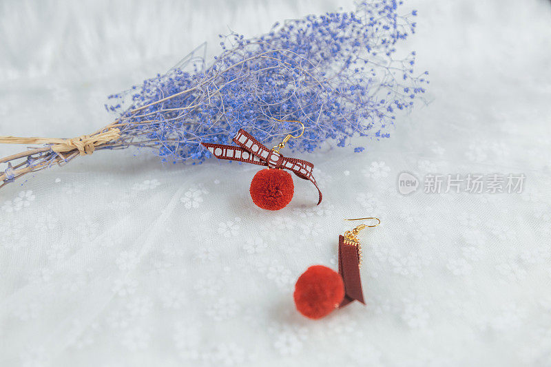 红色的绒球耳环，它的形状是一个毛茸茸的球。一个样品装饰，在耳环旁边有一束蓝色的花。这些东西都放在薄纱上。耳环看起来非常优雅和时尚。