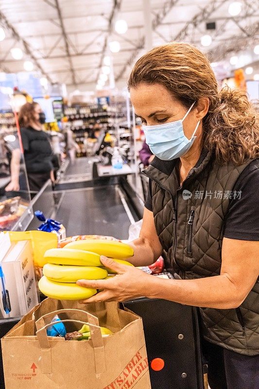 在超市的收银区，带着防护口罩的成熟女性正在收拾她的食品杂货