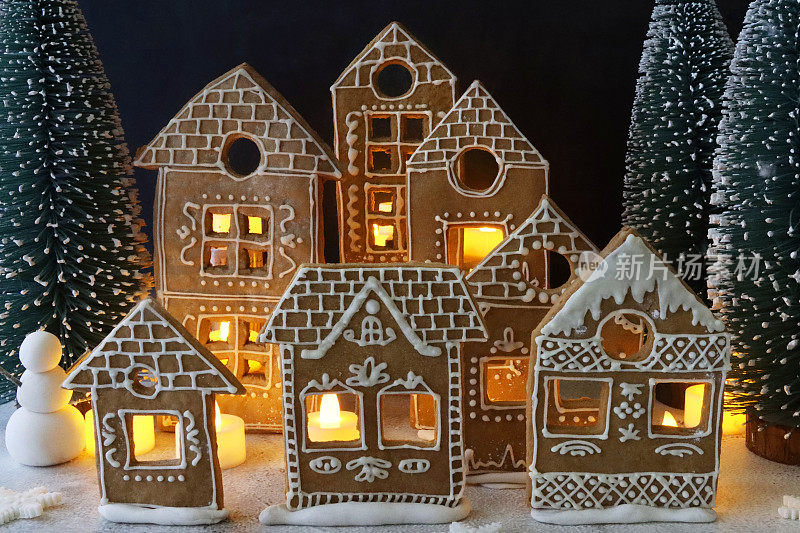 形象自制，姜饼屋饼干装饰白色皇家糖霜展示在雪天，夜晚的圣诞村场景，被仙女灯照亮，雪人，糖霜糖雪，黑色背景