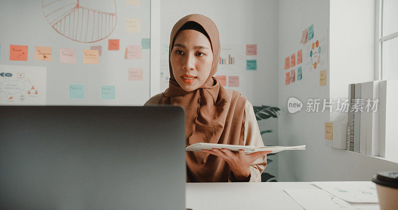 年轻的亚洲穆斯林女商人，戴着希贾布，坐在办公室的办公桌前，通过笔记本电脑和记事本进行视频通话。