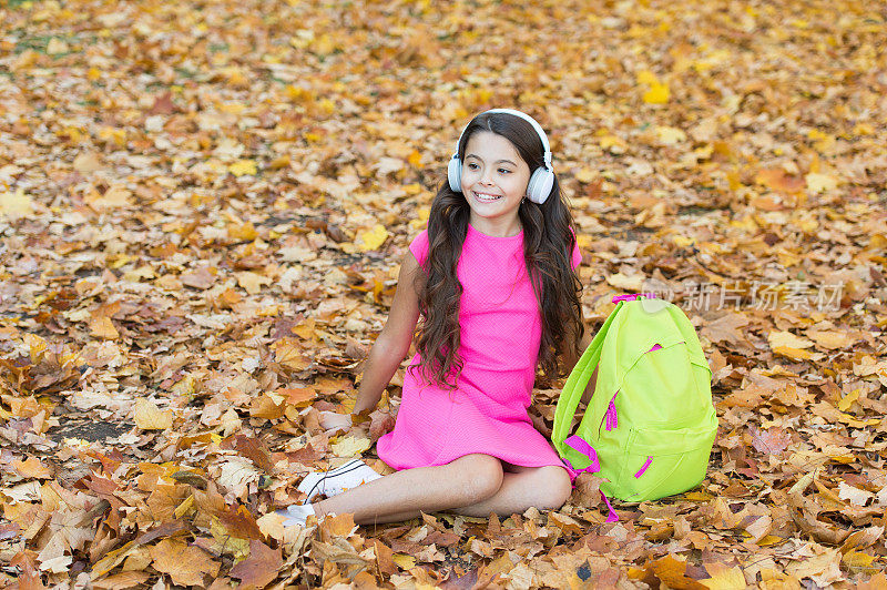 快乐的孩子在秋叶上放松。小女孩戴着耳机。回到学校。现代教育。孩子背着书包在秋天的森林里。知识日的概念。秋天公园里的孩子