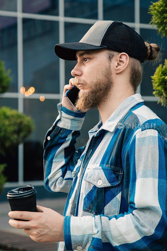 一个留着胡子的年轻人正在讲电话，手里拿着一杯咖啡。