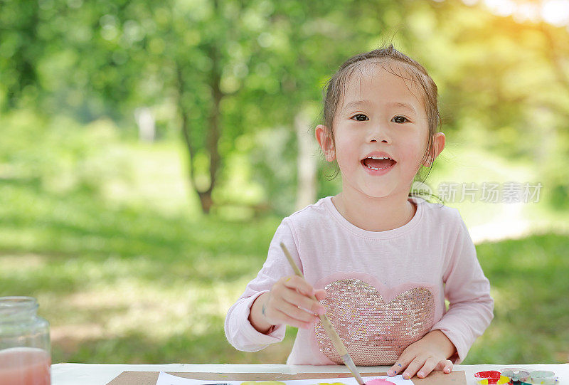 快乐的亚洲儿童女孩坐在一张桌子在一个夏天的花园绘画用画笔。