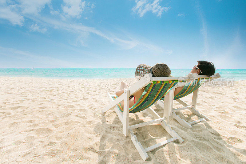 一对夫妇在沙滩椅上晒日光浴海滩是亮蓝色的。在夏天