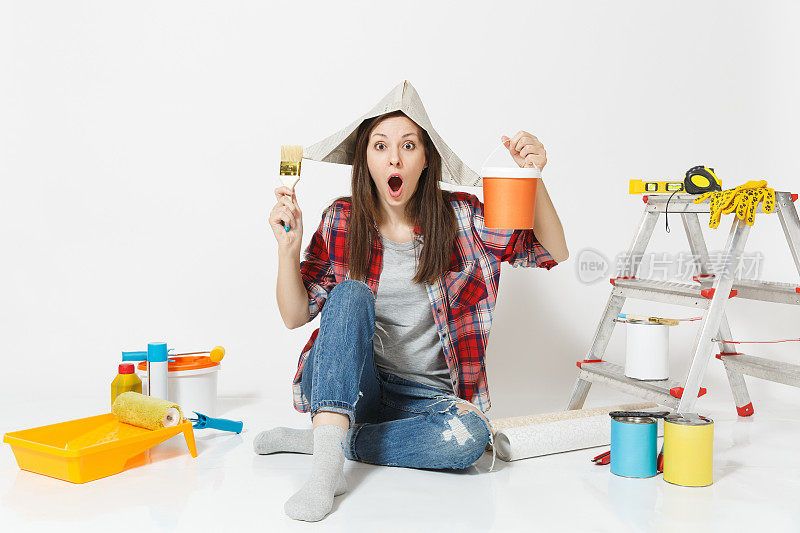 女人戴着报纸帽坐在地板上，用刷子、油漆罐、仪器把公寓装修隔离在白色的背景上。墙纸，胶水配件，油漆工具。修理家里的概念