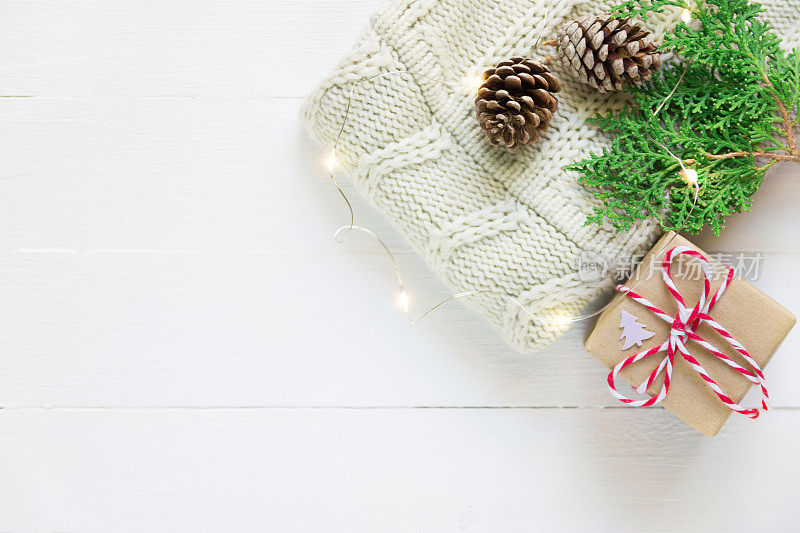 折叠针织灰白色羊毛衫礼盒松果绿杜松小枝金色的灯花环木板桌子靠窗。舒适的冬天的晚上。新年圣诞魔幻气氛。Scandi风格