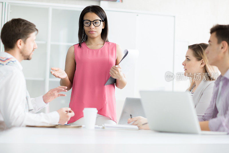 严肃自信的黑人女商业领袖穿着粉红色衬衫站在桌子上，在会议上与专家团队交谈时打手势