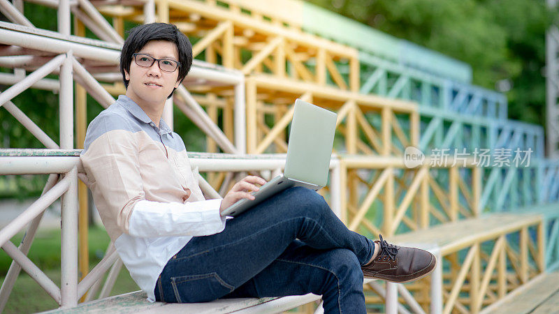 年轻的亚洲男子大学学生使用笔记本电脑站在学院欢呼。校园生活与科技。教育机会或奖学金概念