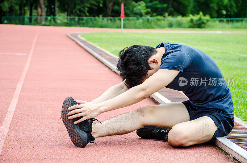 一名男运动员正在热身，一名跑者在跑前坐在体育场的跑道上伸展双腿