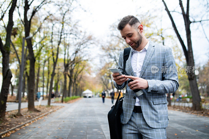 一个商人在公园里走着，拿着一次性咖啡杯看着他的手机。