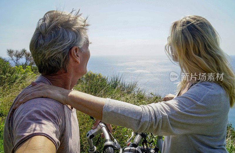 一对成熟的夫妇骑着自行车停下来，在阳光下眺望大海