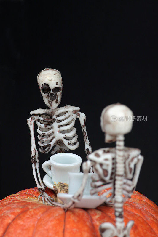 万圣节概念——两个骷髅在黑色背景下的南瓜桌上喝茶