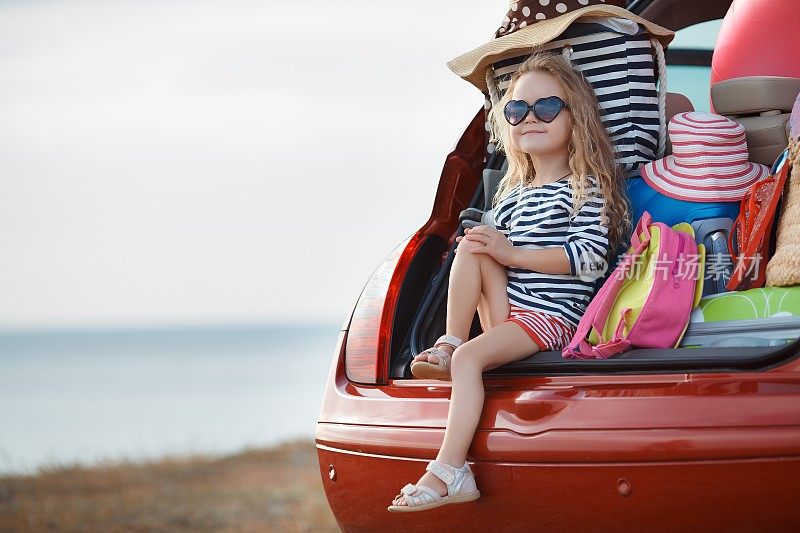 可爱的小女孩坐在装满行李箱的汽车后备箱里，在海边