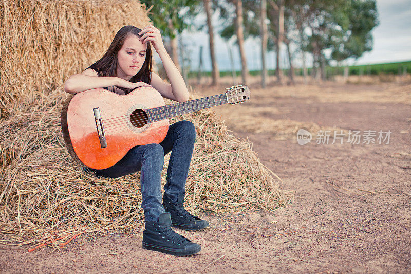 悲伤的年轻女子坐在草堆上，手里拿着原声吉他