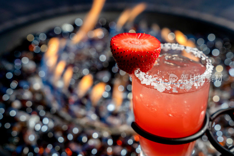 一杯彩色的草莓玛格丽塔，玻璃边镶着盐和草莓装饰，背景是一个丙烷火坑