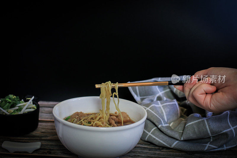 手用筷子夹起美味的鸡蛋面条猪肉和蔬菜在五种香料黑汤在木制桌子上俯视图-亚洲美食风格。