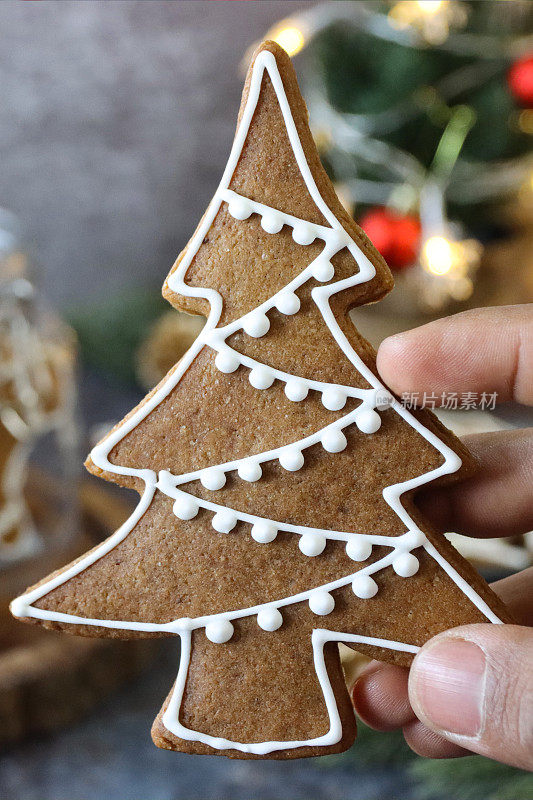一个无法辨认的人拿着一个姜饼圣诞树饼干的特写图像，饼干上装饰着白色的皇家糖霜，玻璃饼干罐里的姜饼人，迷你圣诞树，红色和金色的小装饰物，黑色的背景，家庭烘焙