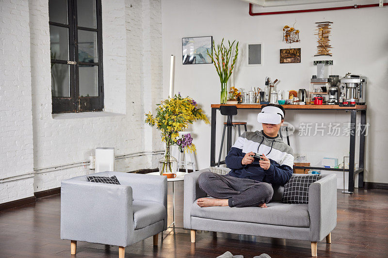 一名东亚男子坐在沙发上享受虚拟现实眼镜。快乐的千禧一代在家里戴着虚拟现实耳机或3D眼镜和手柄玩视频游戏。