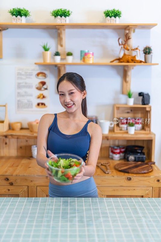 照片年轻美丽的亚洲女性女士在运动服高兴地炫耀一碗沙拉与樱桃番茄在她的房子的厨房节食和减肥健康的生活方式