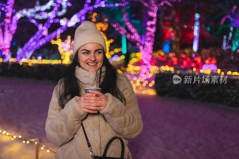 快乐的年轻女子一边喝咖啡一边享受户外的圣诞彩灯