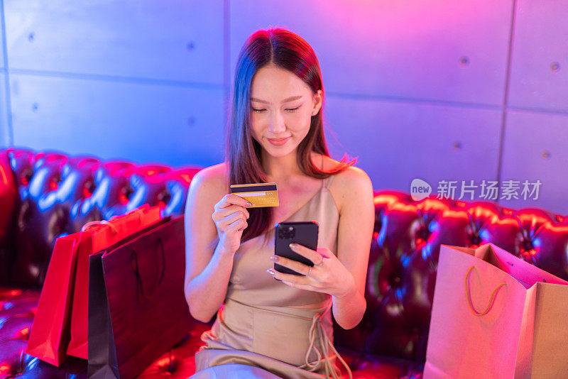肖像照片的年轻美丽的亚洲女模特穿着优雅的衣服在霓虹灯的房间支付与她的信用卡借记卡在她的手机网上购物狂欢
