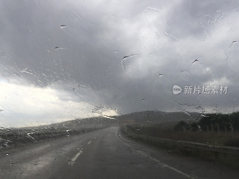下雨天开车。雨点落在汽车挡风玻璃上，多云的天空和空旷的道路背景