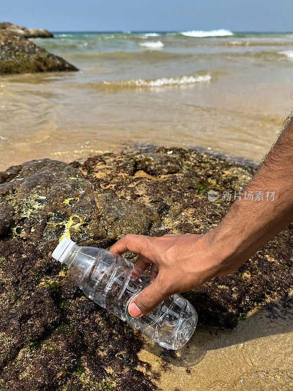图片:在退潮时，白色盖子的透明塑料水瓶被冲走在岩石池中，被不认识的人捡起，海岸上的海洋垃圾和污染，乱扔的沙子，肮脏的海滩，重点在前景