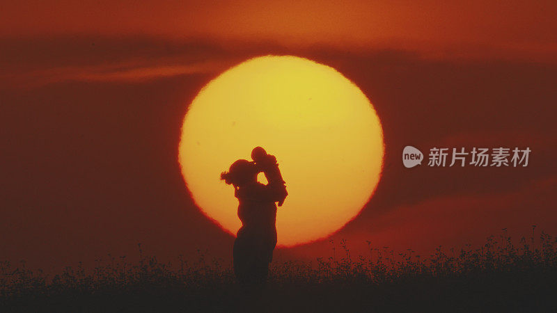 在夕阳的背景下，太阳映衬着橙色的天空，嬉戏的母亲亲吻着婴儿