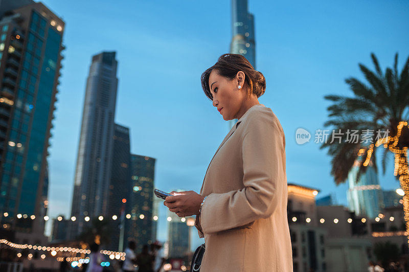 身穿米色西装的亚洲女商人，晚上在市中心用手机发短信的低角度侧视图