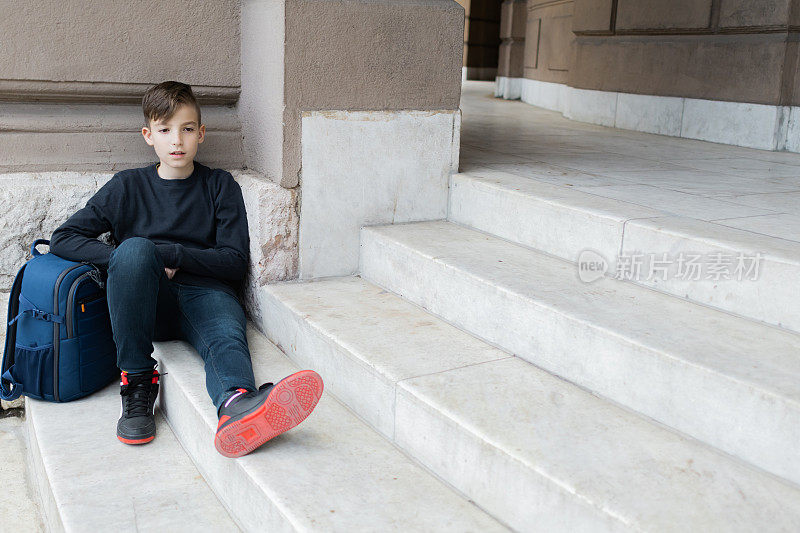 第一天上学，一个背着书包的男生坐在学校大楼附近的楼梯上。回到学校。教育。