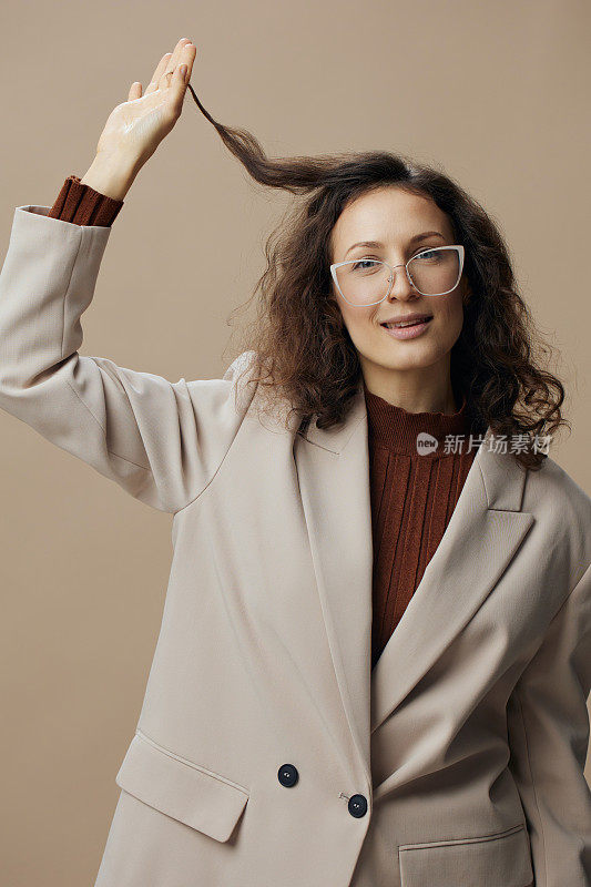 调情开朗可爱快乐快乐的卷曲女性在夹克眼镜触摸头发卷曲看着相机享受好心情在米色背景上摆姿势。酷时尚的眼镜品牌报价