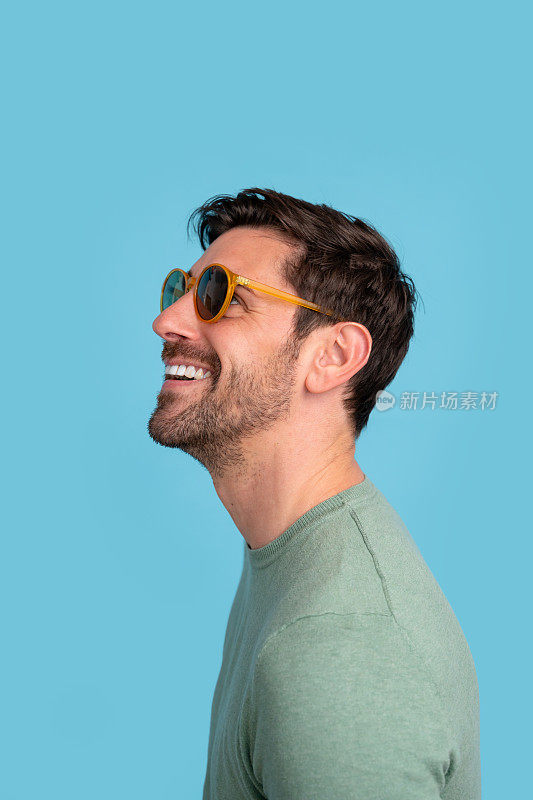 一个快乐的大胡子男人的侧面，他戴着黄色的太阳镜，在舒缓的蓝色背景下，体现了快乐和休闲的风格