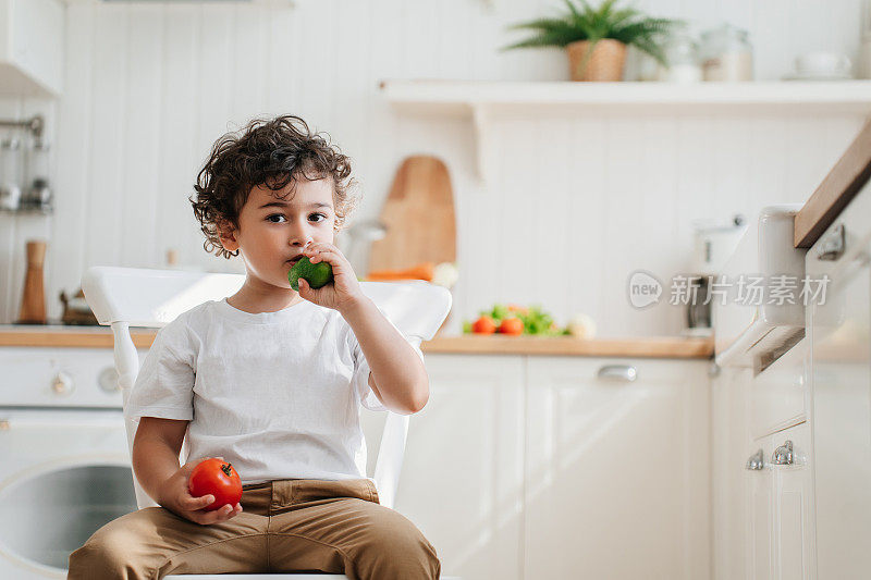 可爱的白人男孩在裤子吃黄瓜拿着西红柿看一边。健康食品的概念。卷曲的西班牙帅哥在厨房吃蔬菜。儿童，学龄前儿童，成长过程。健康饮食