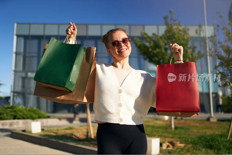 快乐的女人带着购物袋从商场走在城市里。兴奋的女孩享受黑色星期五大减价。女性拿着满满的纸袋在镜头前摆姿势。消费主义的概念。
