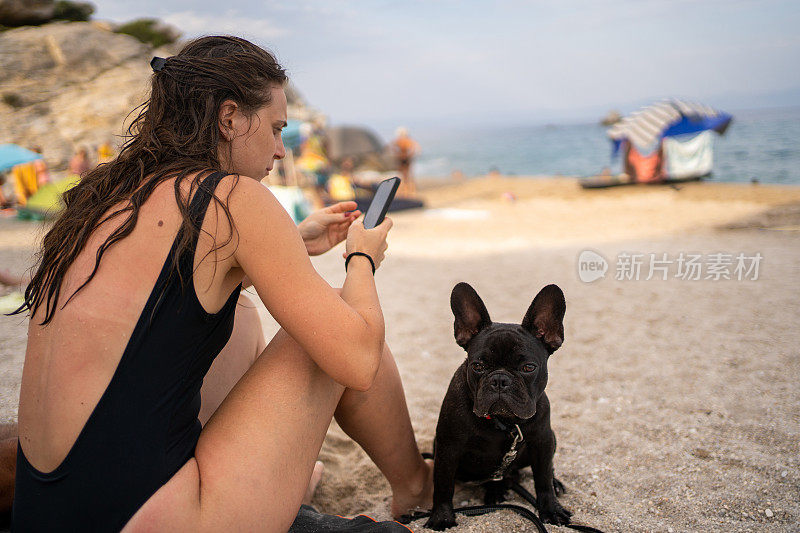 在海滩上，法国斗牛犬坐在年轻女子旁边