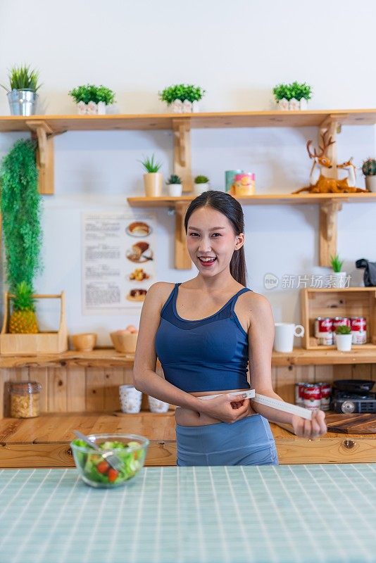 照片的年轻美丽的亚洲妇女女士穿着运动服快乐做腰测量检查她的训练，锻炼和饮食的结果。她家厨房桌上的樱桃番茄沙拉是为了减肥和健康的生活方式