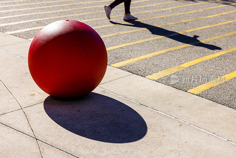 红色的，圆形的，混凝土球体在停车场里，有一个人的影子，沿着马路走着。
