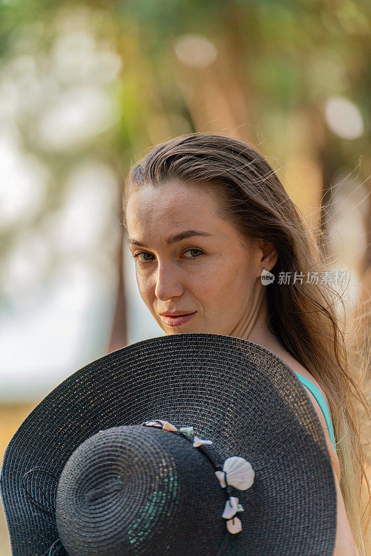 美丽的白人女子在比基尼和黑色沙滩帽摆姿势，在海滩上郁郁葱葱的热带公园快乐地游荡