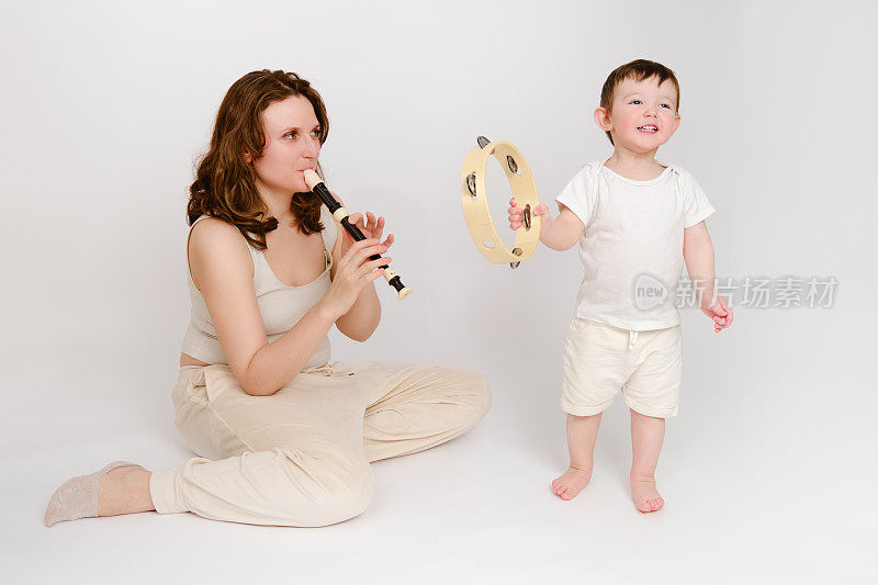 快乐的宝宝和妈妈在工作室白色背景上演奏乐器。一个微笑的孩子与妈妈和吹笛子的肖像。孩子2岁左右(1岁9个月)