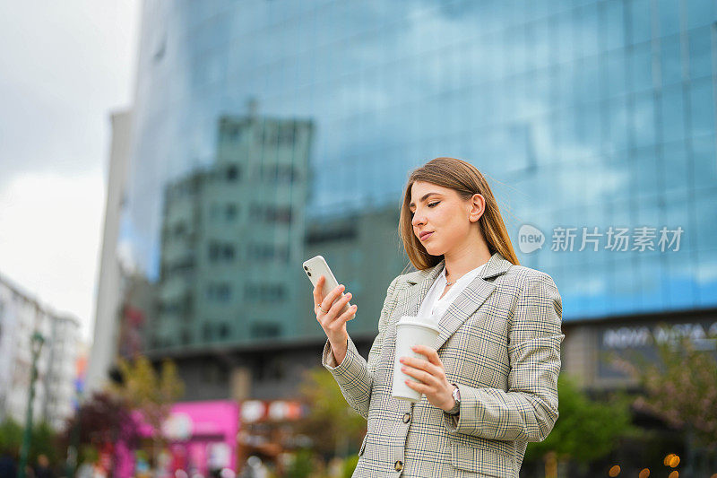 在商务中心前，年轻的女商人拿着咖啡杯看着她的手机