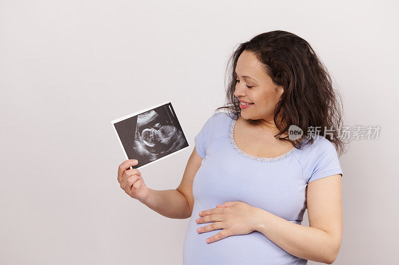 一个快乐的孕妇抚摸腹部的肖像，抱着超声波成像仪哦，她的孩子，隔离在白色背景上