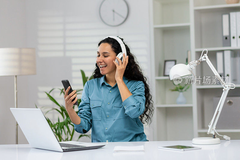 拉丁美洲女人，一头卷发，在家里的工作空间里通过连接智能手机的无线耳机听音乐。兴奋的数字设计师打开鼓舞人心的播放列表，提高工作效率