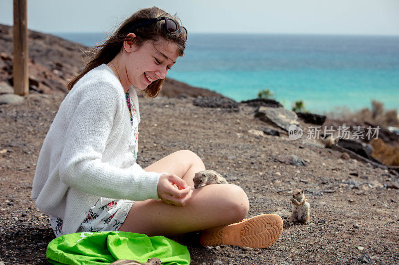 美丽的年轻女孩在富埃特文图拉山区喂一只野生松鼠。漂亮时髦的年轻女孩正在驯养一只野生松鼠。