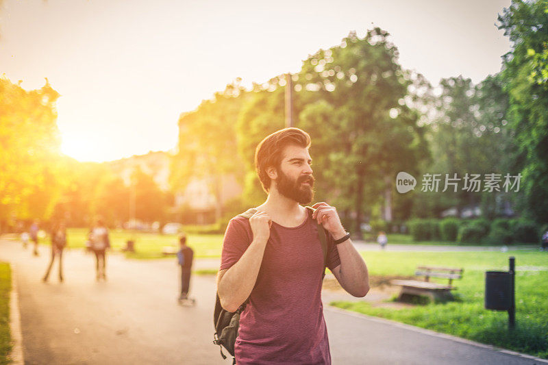 在一个阳光明媚的夏日，一个背着背包的帅气胡子男人在公园里